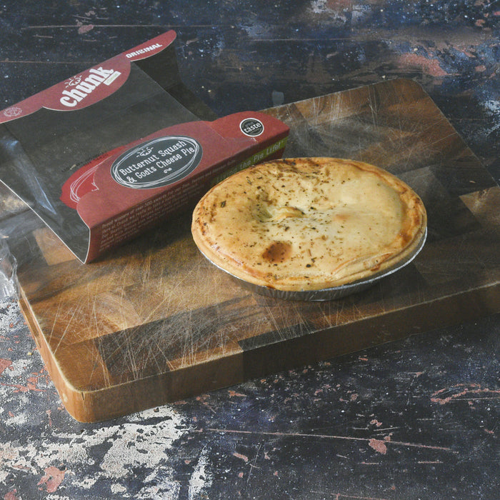 A baked veggie pie sits on a wooden cutting board next to its red and black Chucky branded packaging box, set against a dark rustic background.