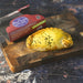 A baked Chunk original butter chicken curry pie sits on a wooden cutting board next to its red and blue packaging box. The pie has a golden-brown crust and is sprinkled with black nigella seeds.