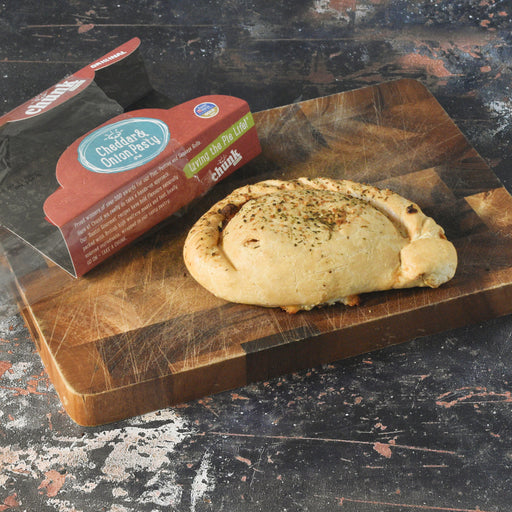 A baked cheese and onion pastry sits on a wooden cutting board next to its branded retail packaging, photographed on a dark rustic tabletop.