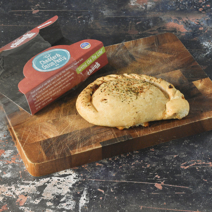 A baked cheese and onion pastry sits on a wooden cutting board next to its branded retail packaging, photographed on a dark rustic tabletop.