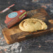 A baked cheese and onion pastry sits on a wooden cutting board next to its branded retail packaging, photographed on a dark rustic tabletop.