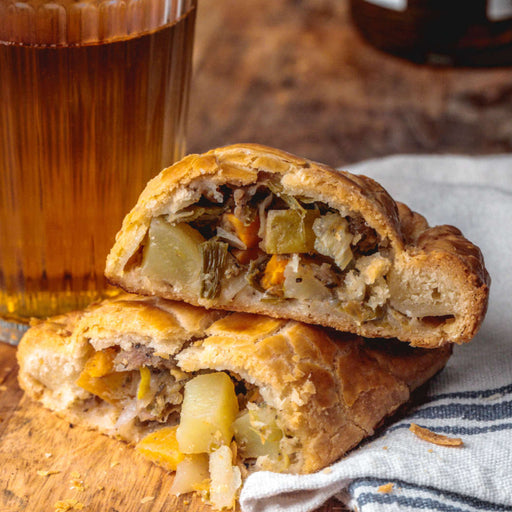 Chunk original Pork & Scrumpy pasty cut open and placed on cloth a wooden cutting board with a glass of cide to one side.