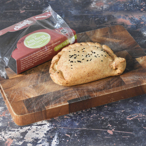 A Chunk Original baked handmade pasty with black sesame seeds sits on a wooden cutting board next to its branded retail packaging box. The box is wrapped in clear plastic and features a dark red label with a green circular logo.