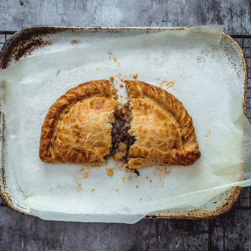 Chunk original Steak pasty, baked on a baking tray, cut in half with filling spilling out, on a rustic metal surface.