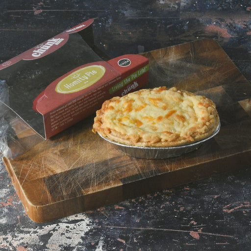 A baked Chunk Homity pie sits in a foil pan on a wooden cutting board, with its red and black retail box placed behind it on a dark rustic tabletop.