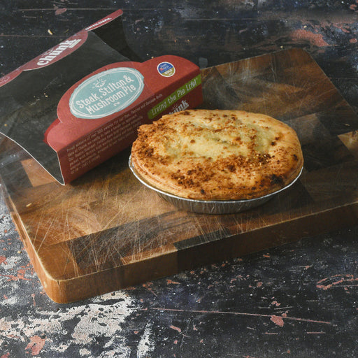 A Chunk baked steak and mushroom pie in a foil tin sits on a wooden cutting board next to its red and black retail packaging box.