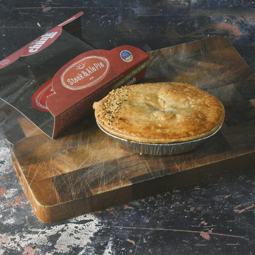 A baked Chunk Original Steak & Pie pie in a foil tin sits on a wooden cutting board next to its branded cardboard packaging box.