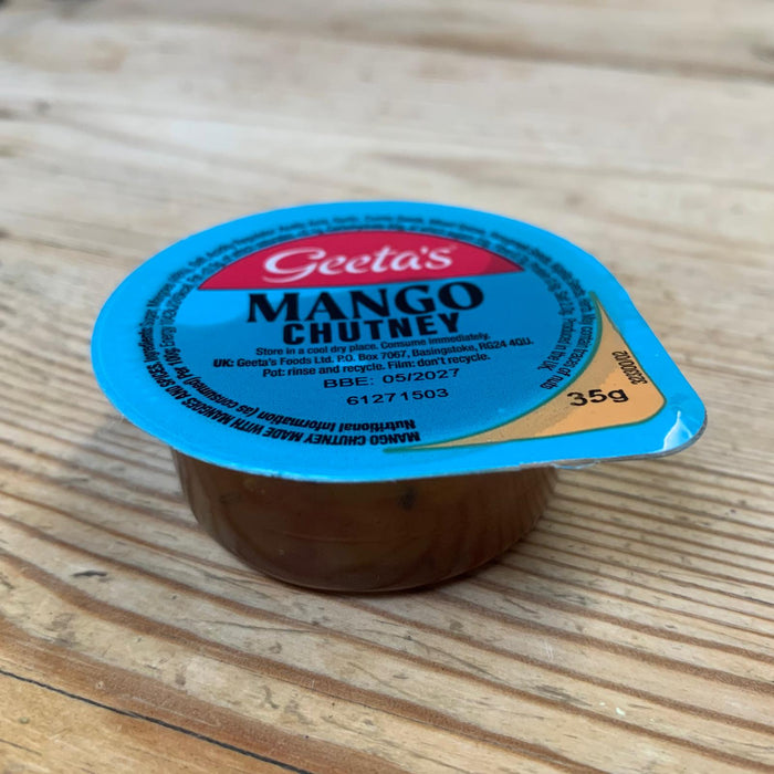 A small Geeta's Mango Chutney container with a bright blue lid sits on a light wood grain surface.